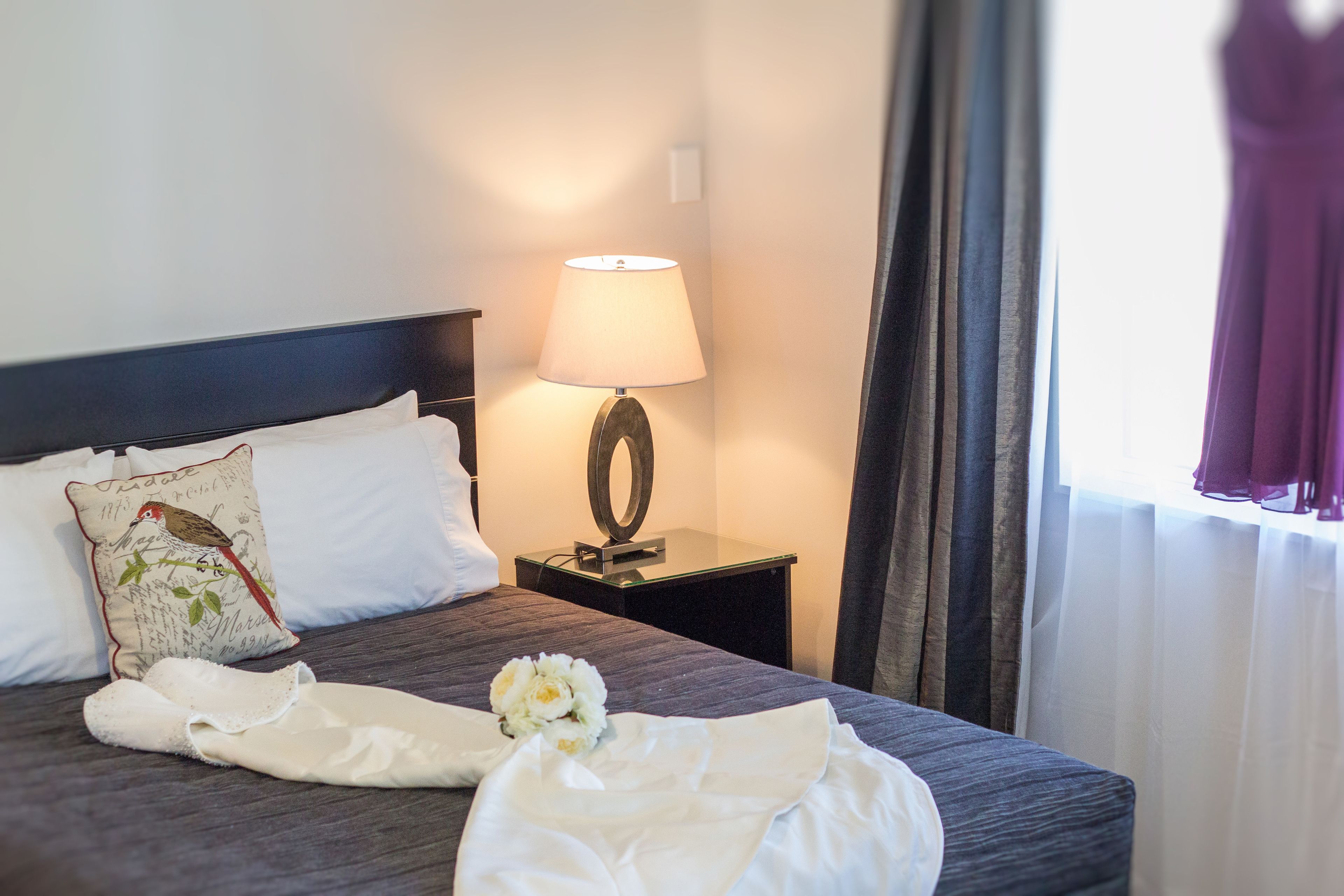 A neatly made bed with white linens, a decorative pillow, a bedside lamp, and a bouquet of flowers.