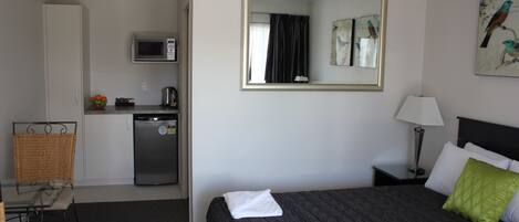 A hotel room with a bed, a small kitchenette, a microwave, a refrigerator, a chair, a nightstand with a lamp, and a painting on the wall.