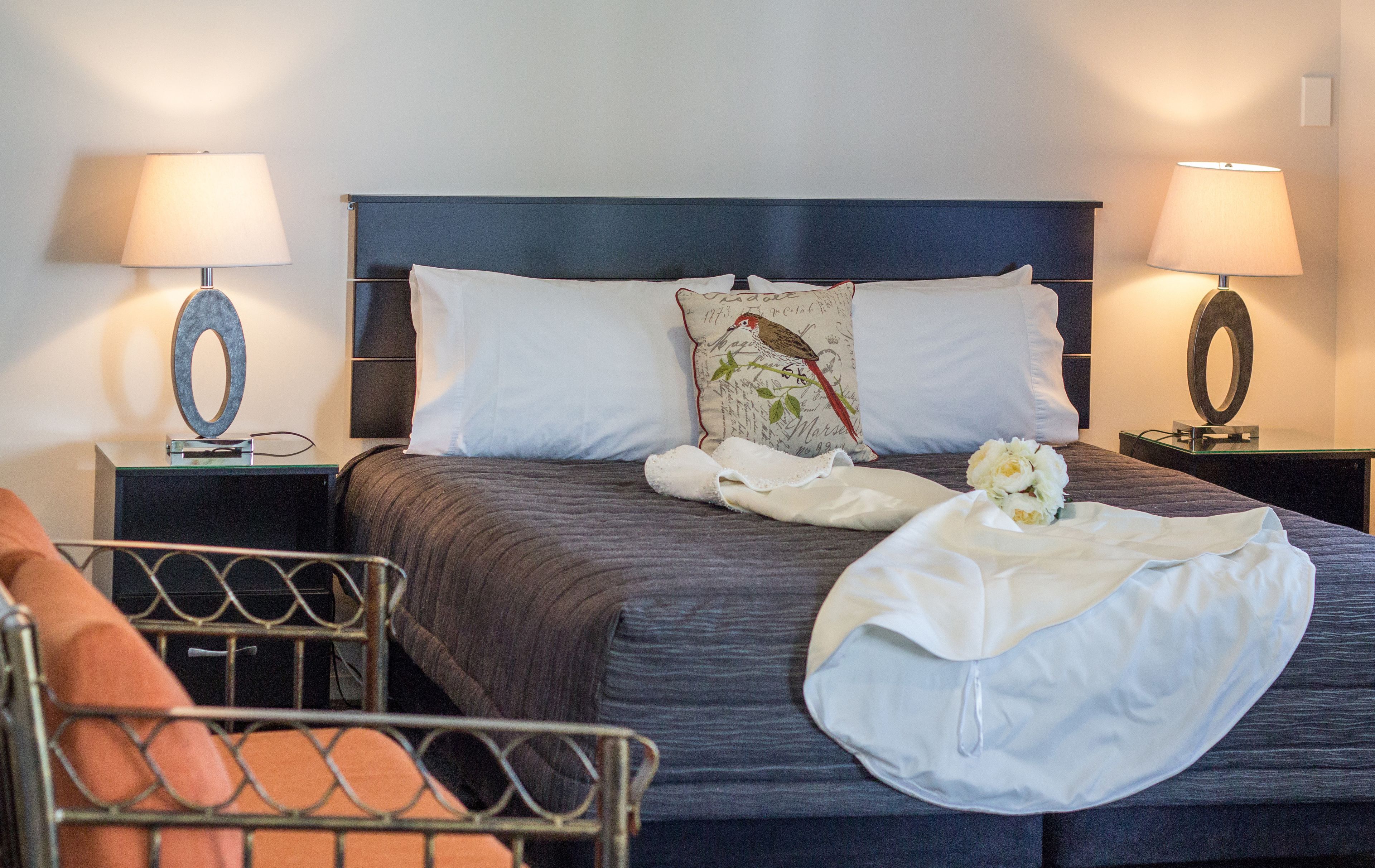 A neatly made bed with a dark headboard, white pillows, and a decorative pillow with a bird design. There are two bedside lamps with circular bases and a small table with a lamp. A white cloth and a bouquet of flowers are on the bed.