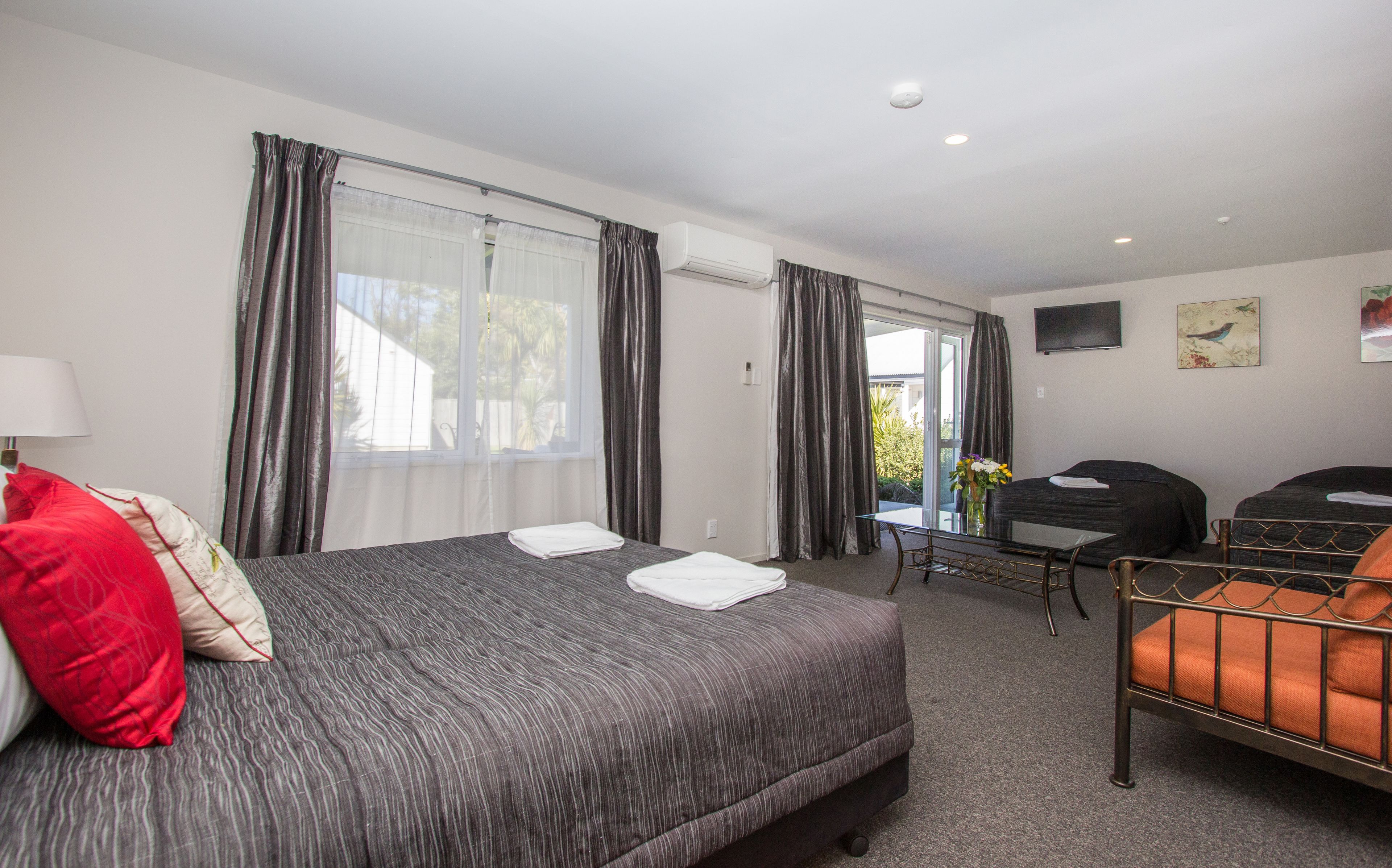 A hotel room with a large bed, a desk, and a chair. There is a window with curtains, an air conditioning unit, and a flat-screen TV on the wall.