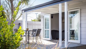A covered patio with a table and chairs, a sliding glass door leading to a room, and a view of a fence and trees. - Greyfriars Motel (Greytown)