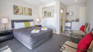 A hotel room with a large bed, bedside tables, a glass dining table, and a kitchenette. - Greyfriars Motel (Greytown)