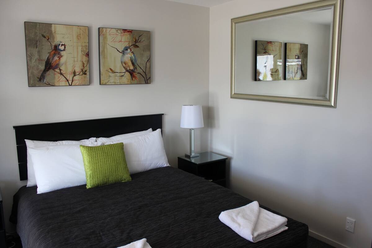 A neatly made bed with white and green pillows, a bedside lamp, and two wall-mounted paintings of birds.