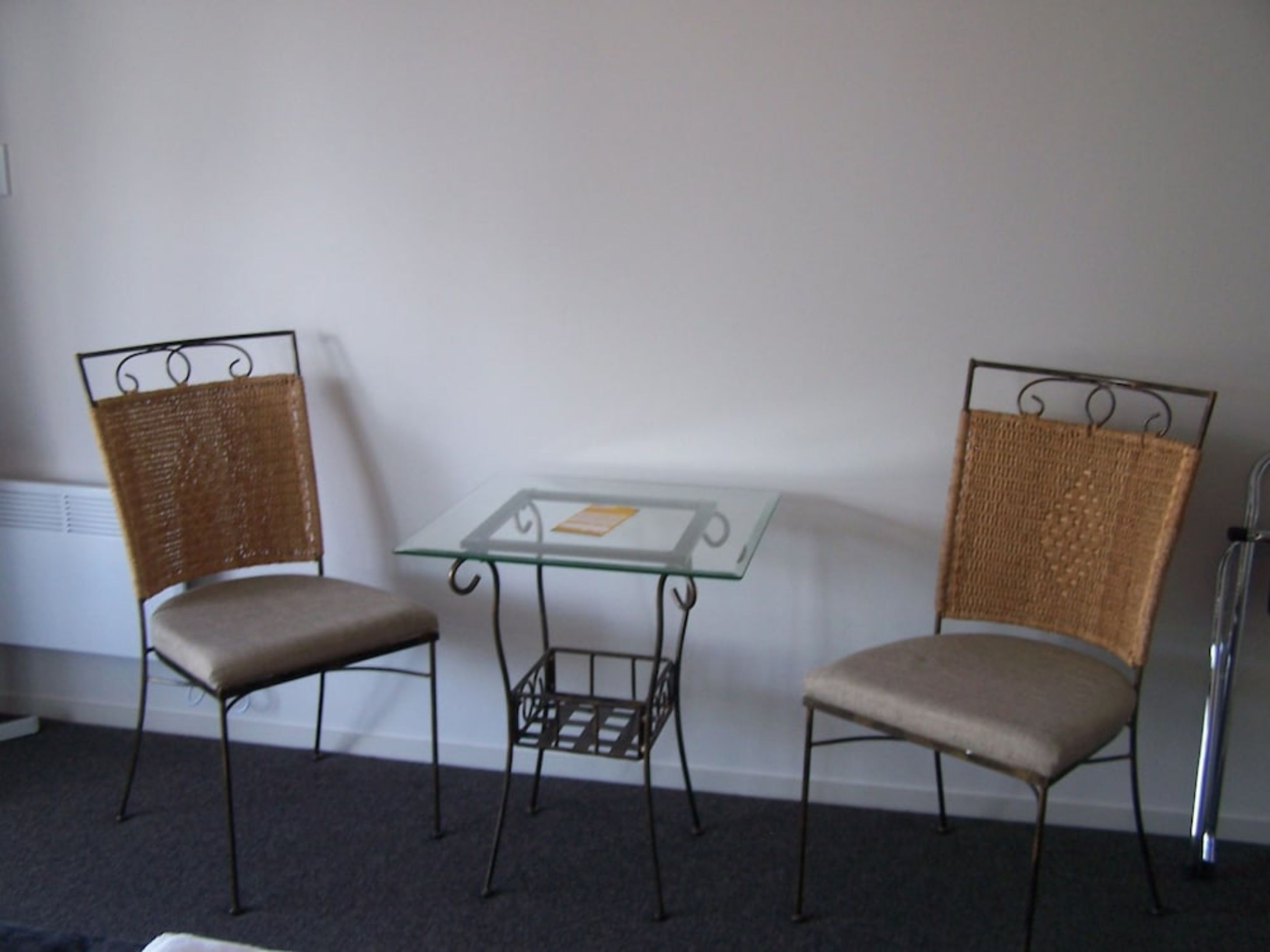 Two wicker chairs with cushioned seats and backs, a small glass table with a metal frame, and a radiator on the wall.