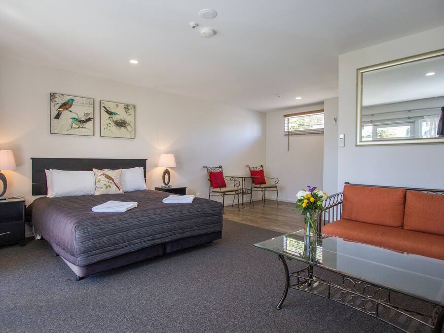 Una habitación de hotel con una cama grande, dos mesitas de noche con lámparas, una mesa de centro de cristal, un sofá, una silla y una obra de arte en la pared.