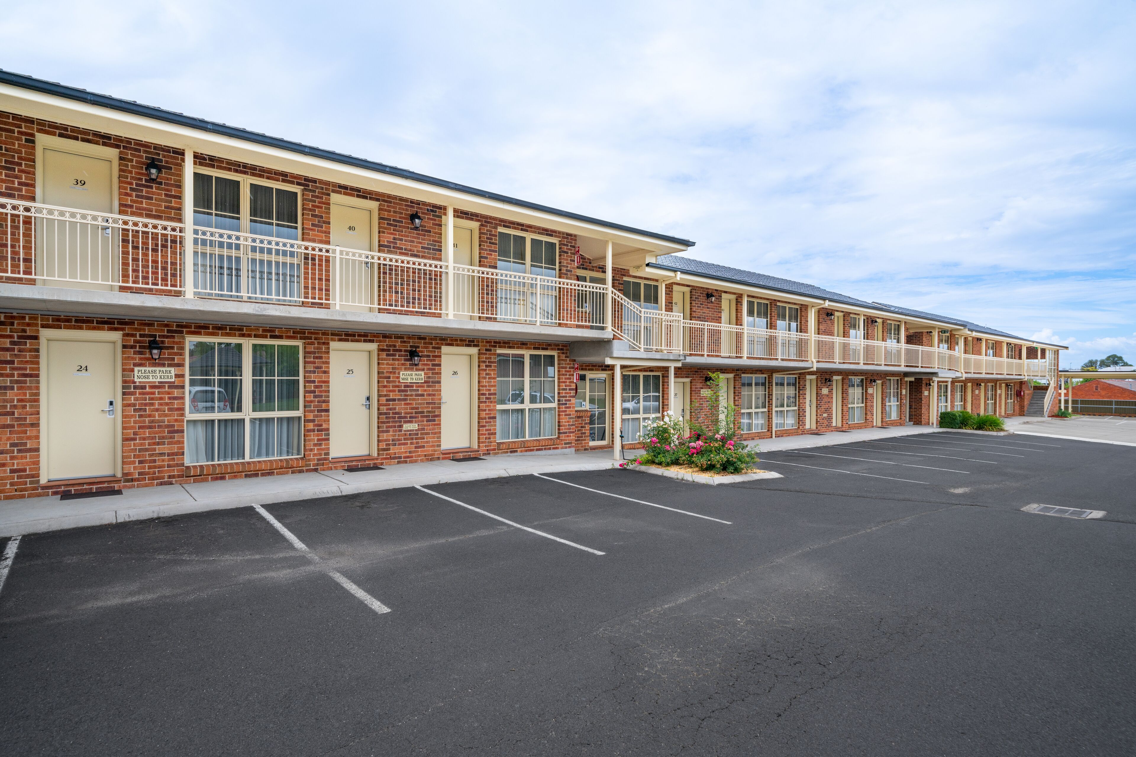 Un motel de varios pisos con habitaciones numeradas, un estacionamiento y un pequeño jardín.