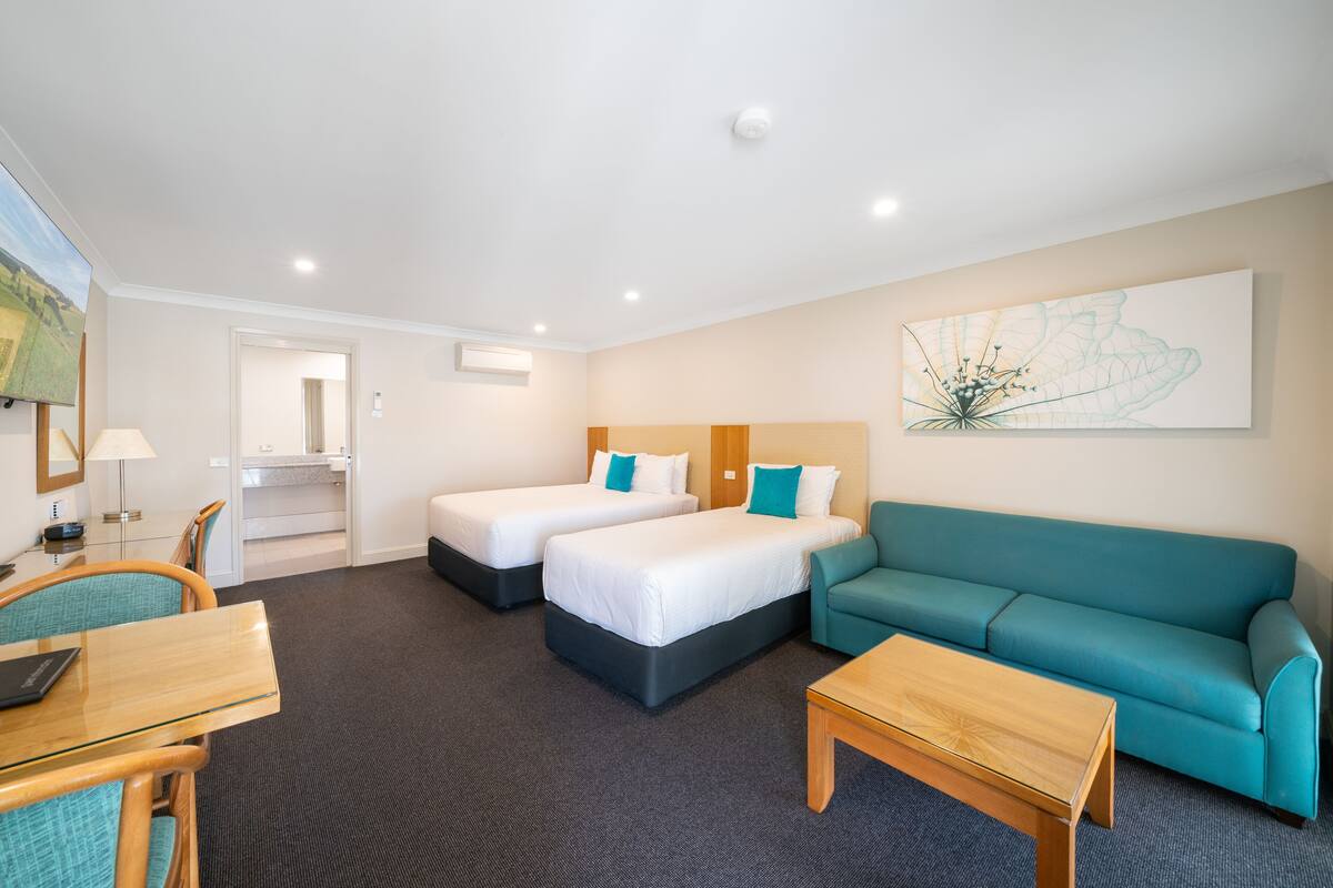 A hotel room with two beds, a teal sofa, a wooden coffee table, and a desk with a lamp.
