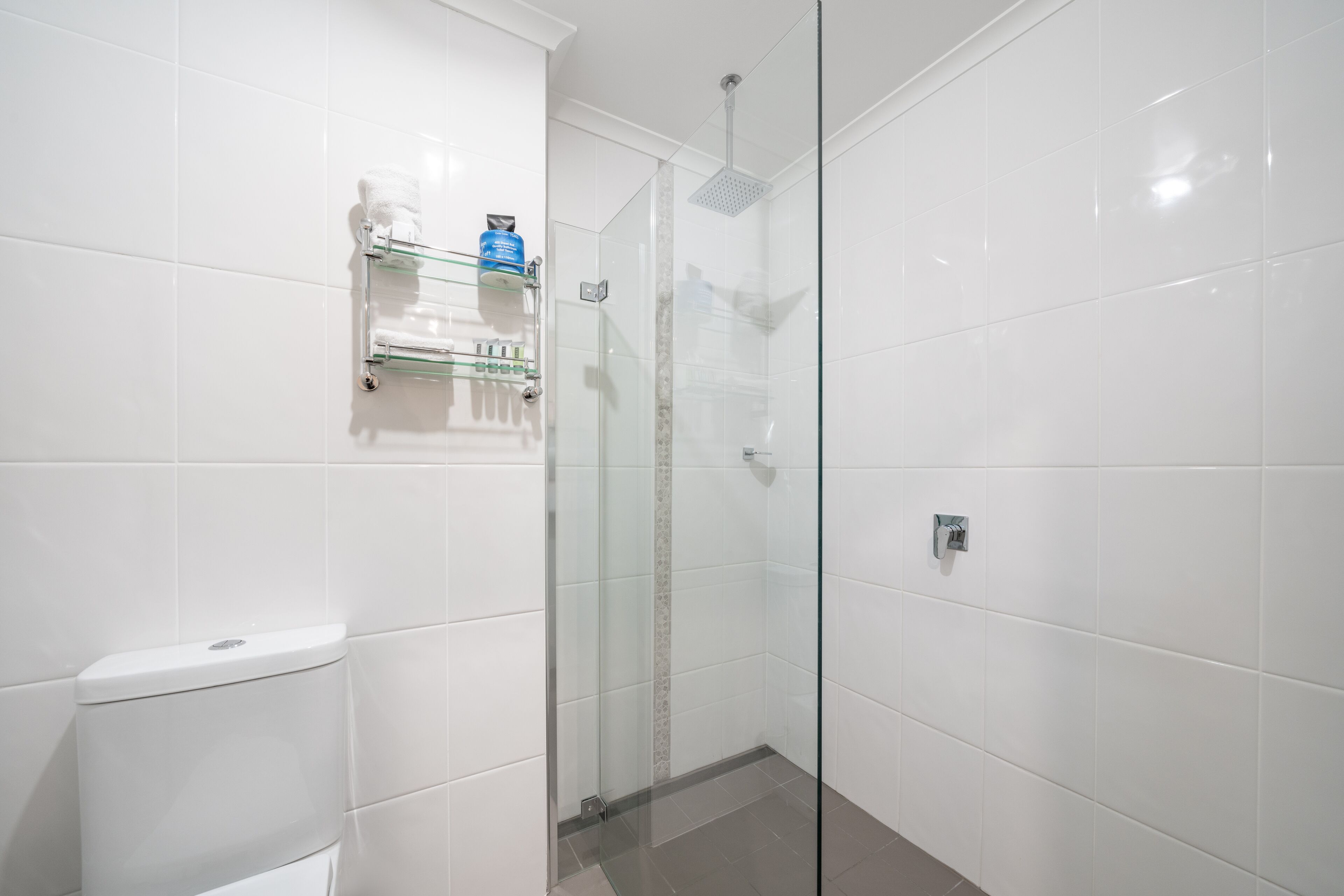 Un baño moderno con azulejos blancos, cabina de ducha de vidrio, inodoro y estante con artículos de aseo.