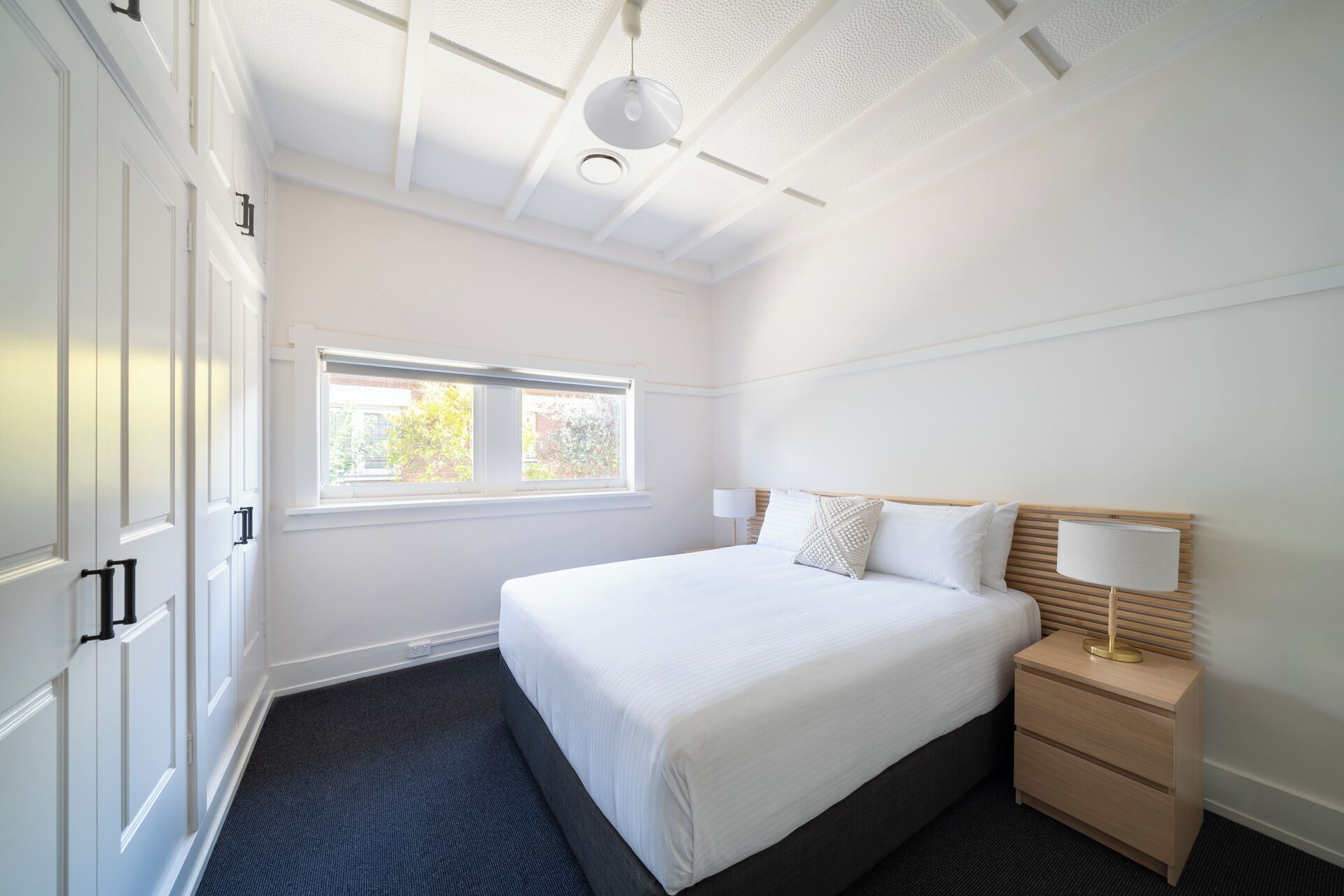 A hotel room with a large bed, a bedside table with a lamp, a window, and a ceiling with recessed lighting.