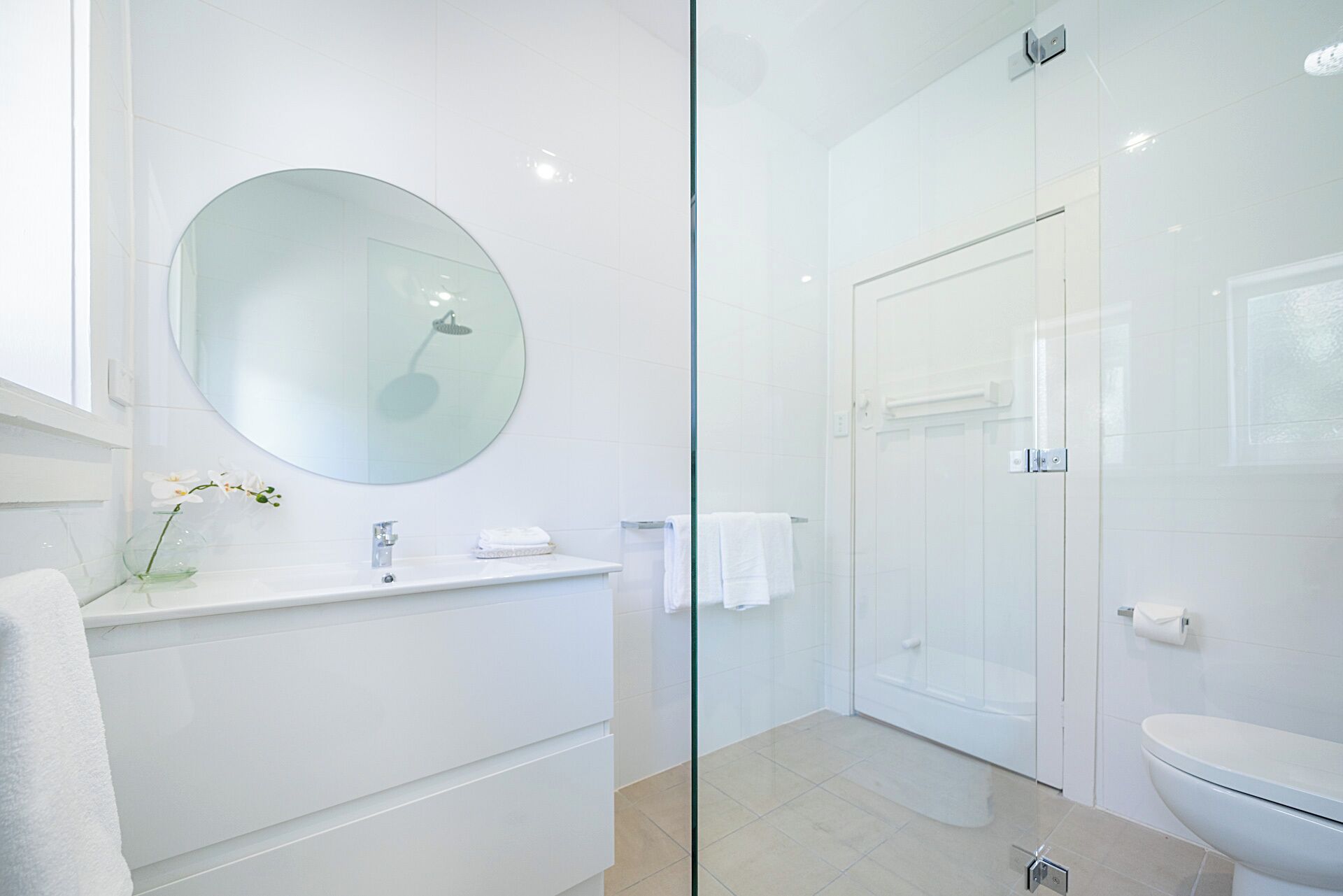 Un bany modern amb dutxa de vidre, un mirall rodó i un lavabo blanc.