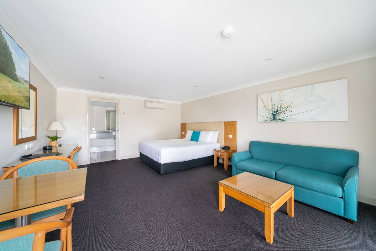 A hotel room with a bed, a teal sofa, a wooden coffee table, a desk with a chair, and a painting on the wall.