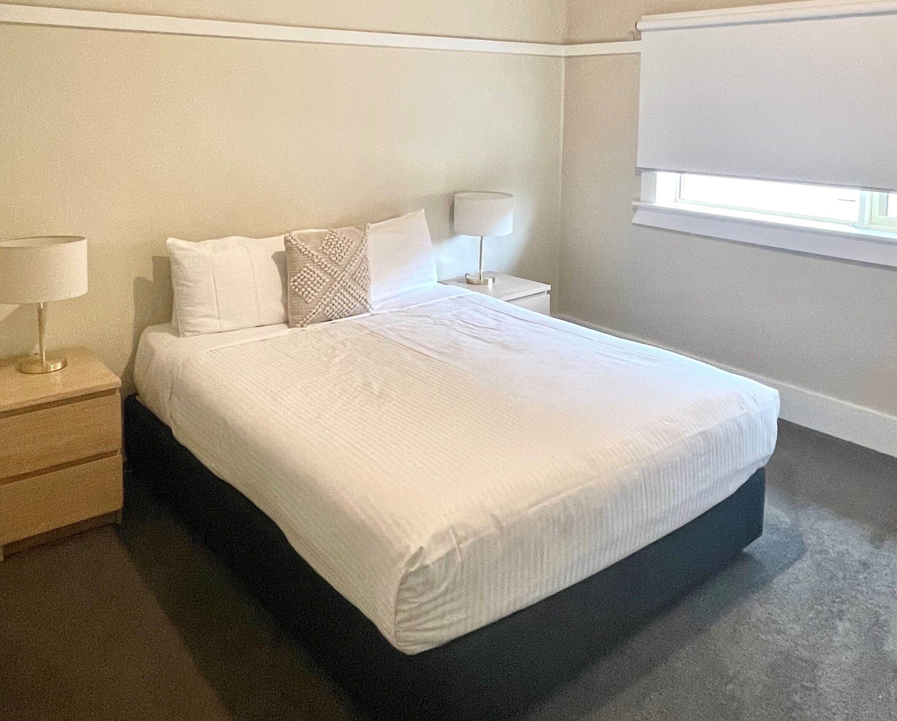 Una cama bien tendida con ropa blanca, una mesita de noche con lámpara y una ventana con persianas.