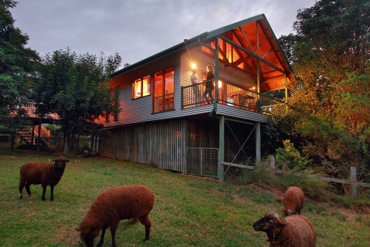 A rustic barn-style house with a balcony, surrounded by a grassy area with sheep.