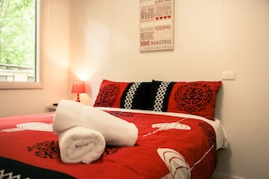 Un lit bien fait, recouvert d’une couvre-lit à motifs rouges et blancs, avec une serviette blanche roulée et des coussins décoratifs aux motifs géométriques.