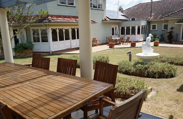A covered patio with a wooden table and chairs, a white fountain, and a garden area with potted plants. - Melville House (Lismore)