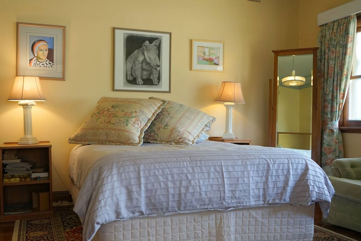 Un lit bien fait, avec une couette blanche matelassée, deux oreillers et une table de chevet assortie, sur laquelle se trouve une lampe. Des œuvres d’art encadrées ornent le mur au-dessus du lit.