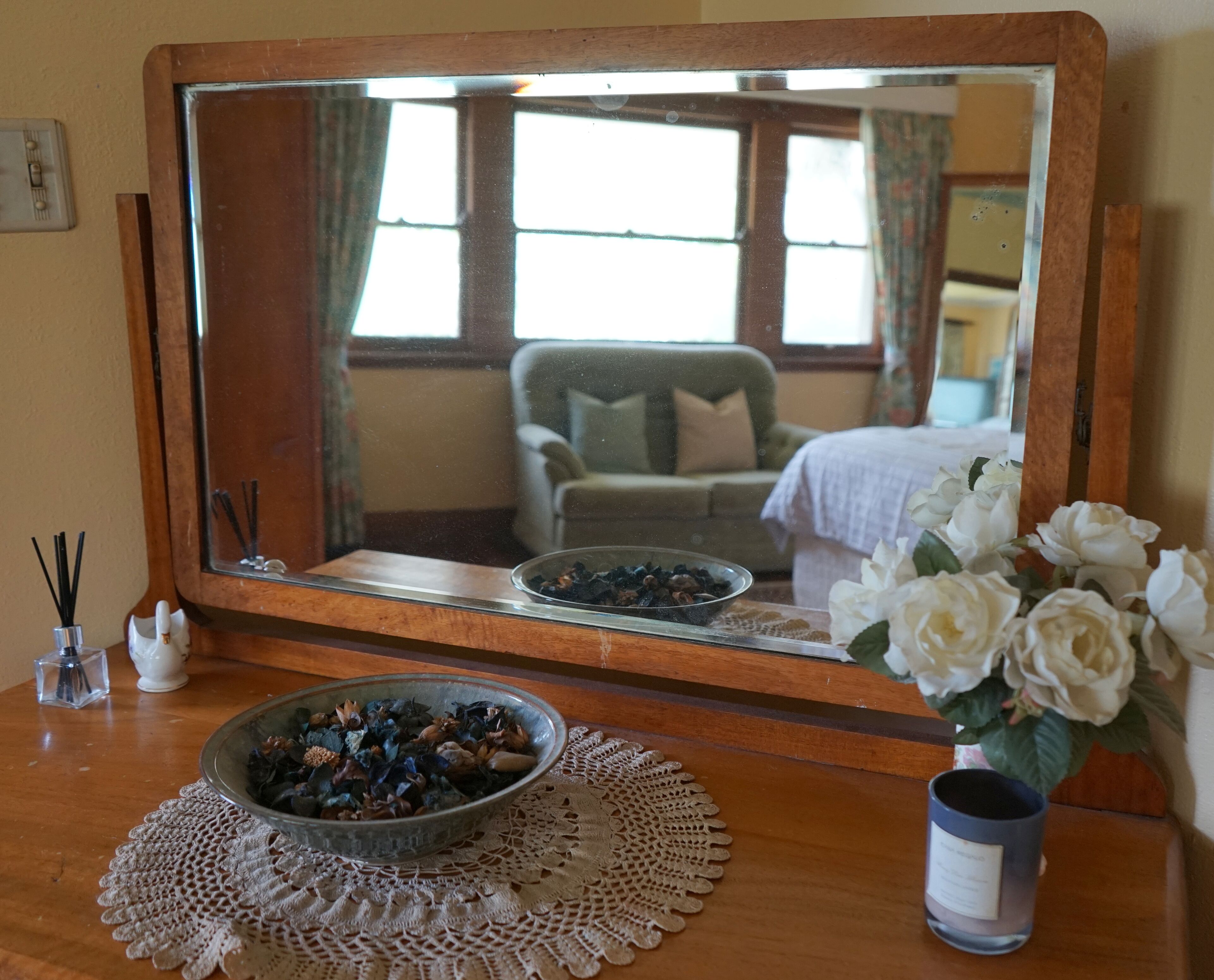 A wooden vanity with a large mirror, a bowl of dried flowers, a vase of white roses, and a candle holder.