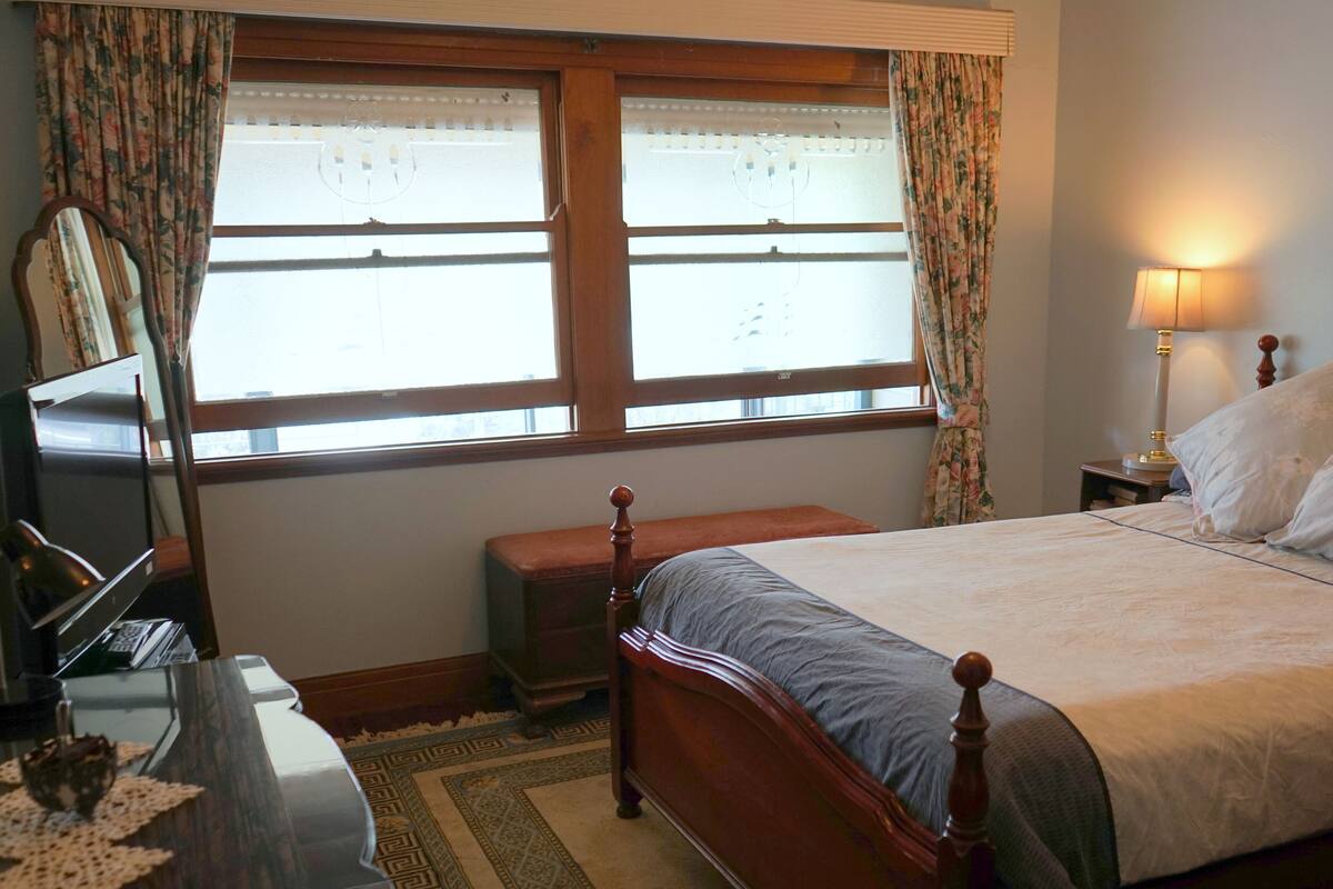 Une chambre à coucher avec un lit en bois, une table de chevet avec une lampe, une commode, un miroir et une fenêtre avec des rideaux.