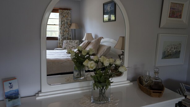 A bedroom with a large mirror, a bedside table with a vase of flowers, and a bed with pillows and a blanket. - Melville House (Lismore)