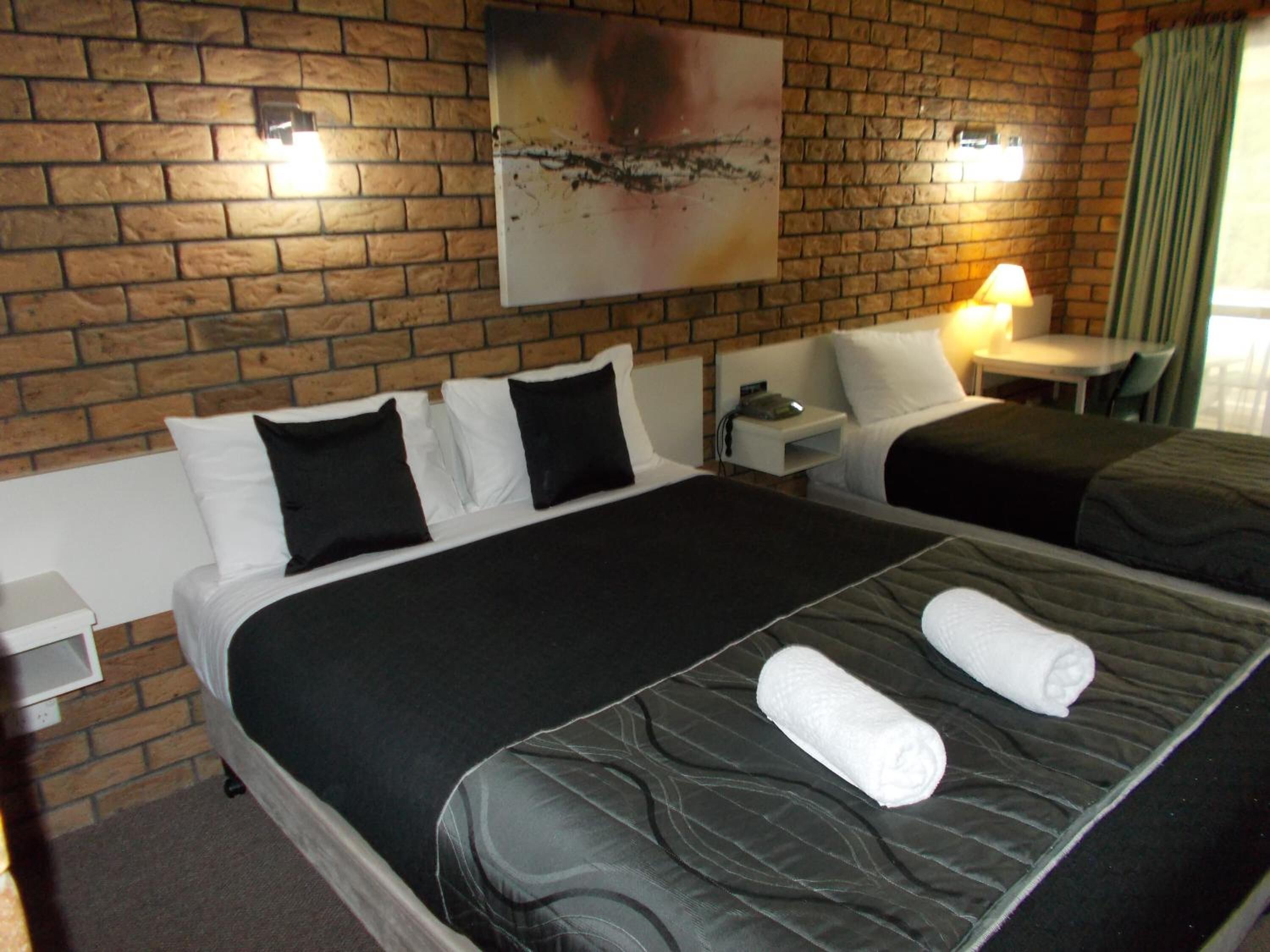 A hotel room with two beds, a brick wall, a painting, a nightstand, and a table with a lamp.