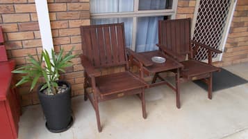 A wooden patio set with a table and four chairs, a potted plant, and a brick wall with windows.