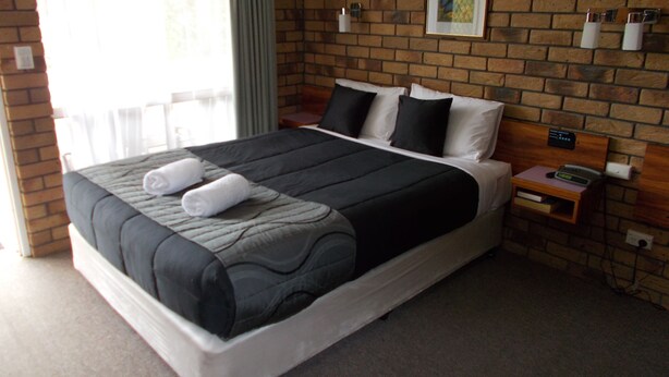 A hotel room with a brick wall, a bed with black and white bedding, a small bedside table with a phone, and a window with curtains. - Park House Motor Inn (Oakey)