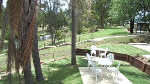A patio with white metal furniture, a small pond, and a grassy area with trees. - Park House Motor Inn (Oakey)