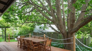 A wooden deck with a table and chairs, surrounded by a rope fence, and a large tree in the background. - Oak Tree Lodge (Rhyll)