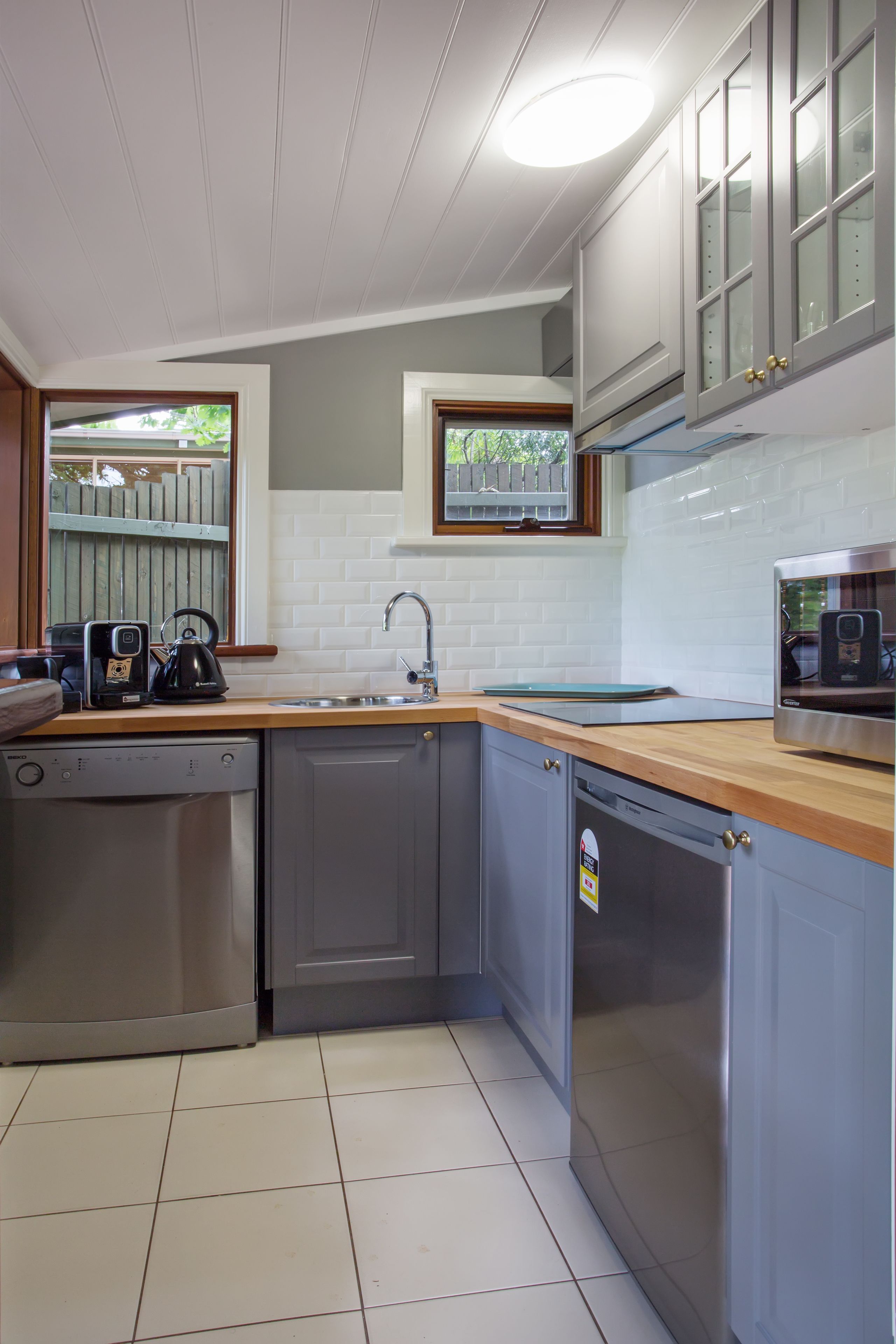 A modern kitchen with grey cabinets, a stainless steel dishwasher, a sink, and a window with a view of greenery.