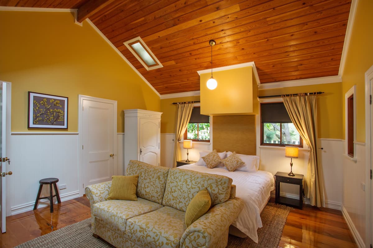 A bedroom with a wooden ceiling, a bed with white linens, a sofa with patterned cushions, and a nightstand with a lamp.