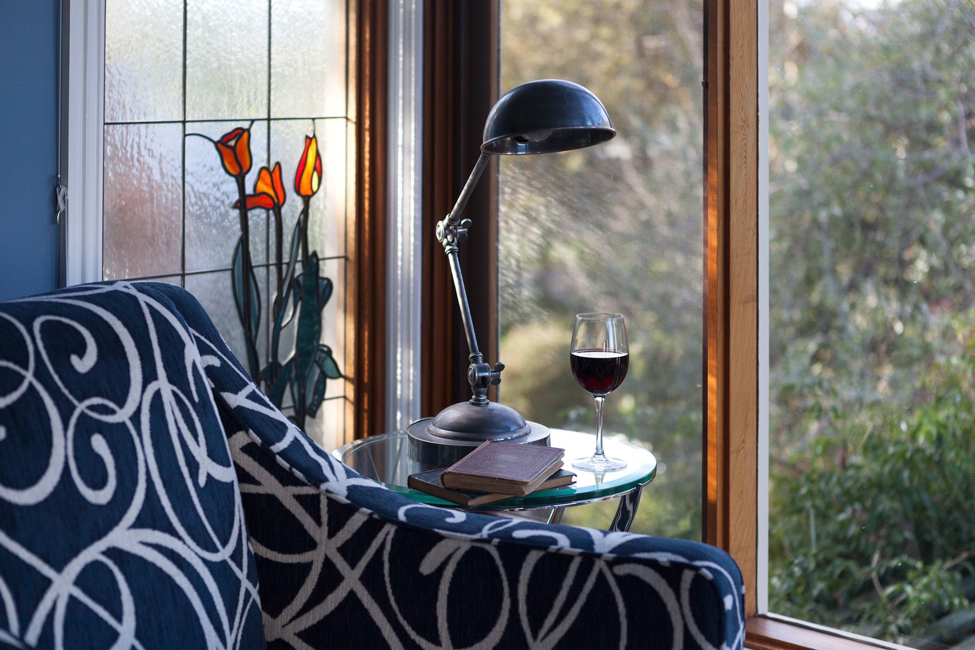 A cozy room with a patterned sofa, a table with a book and a glass of red wine, a lamp, and a window with a view of greenery.