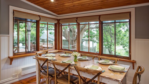 A dining area with a wooden table set for six, surrounded by wooden chairs, large windows with white frames, and a wooden ceiling. - Oak Tree Lodge (Rhyll)