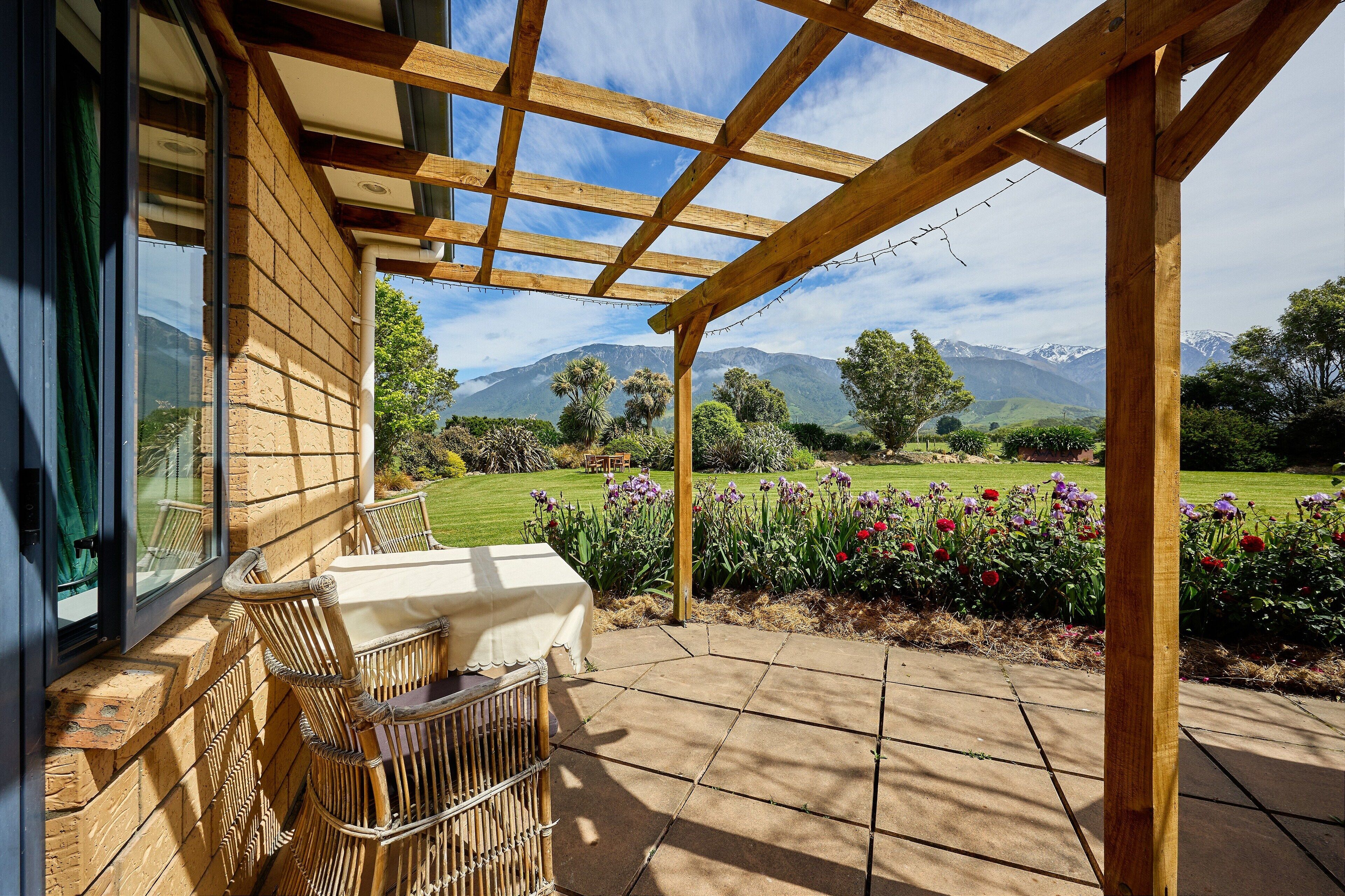 A covered patio with a wooden pergola, a table with chairs, and a view of a landscaped garden with flowers and distant mountains.