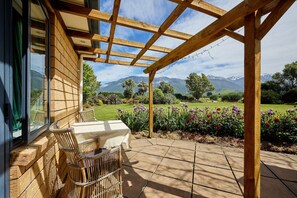 A covered patio with a wooden pergola, a table with chairs, and a view of a landscaped garden with flowers and distant mountains.
