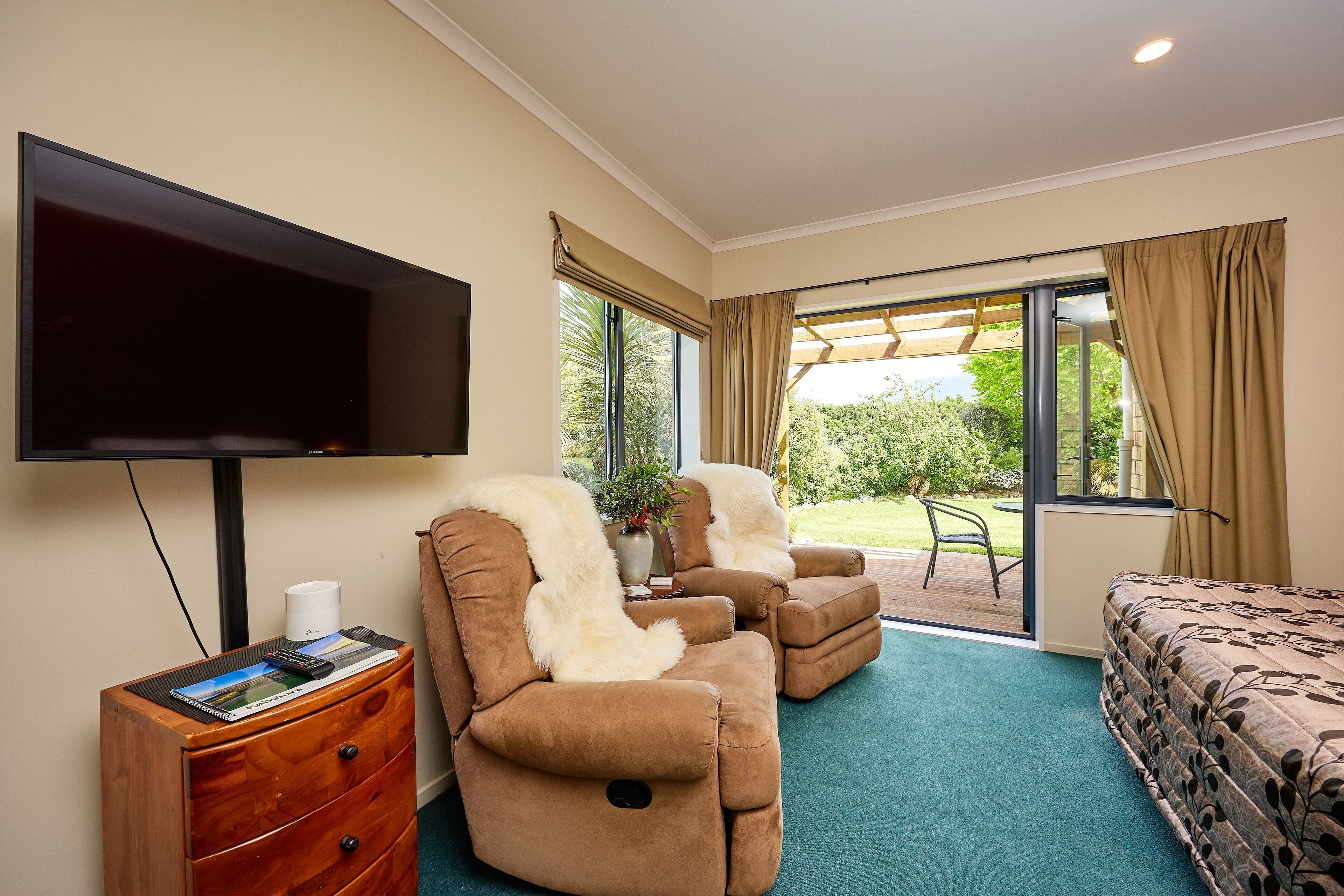 A hotel room with a flat-screen TV, a wooden dresser, a sofa with a white fur throw, a bed with a leaf-patterned duvet, and a sliding glass door leading to an outdoor area.
