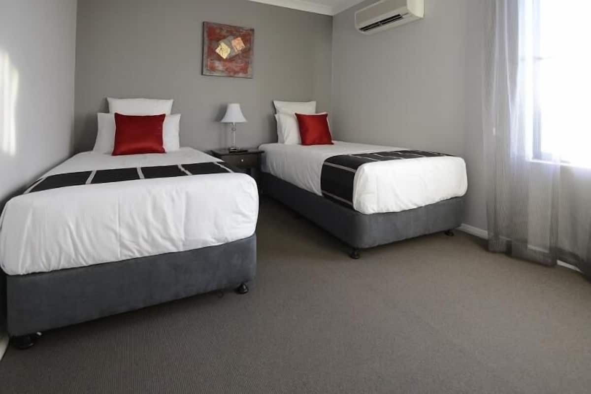 A hotel room with two beds, each with white bedding and red pillows, a bedside table with a lamp, and a painting on the wall.