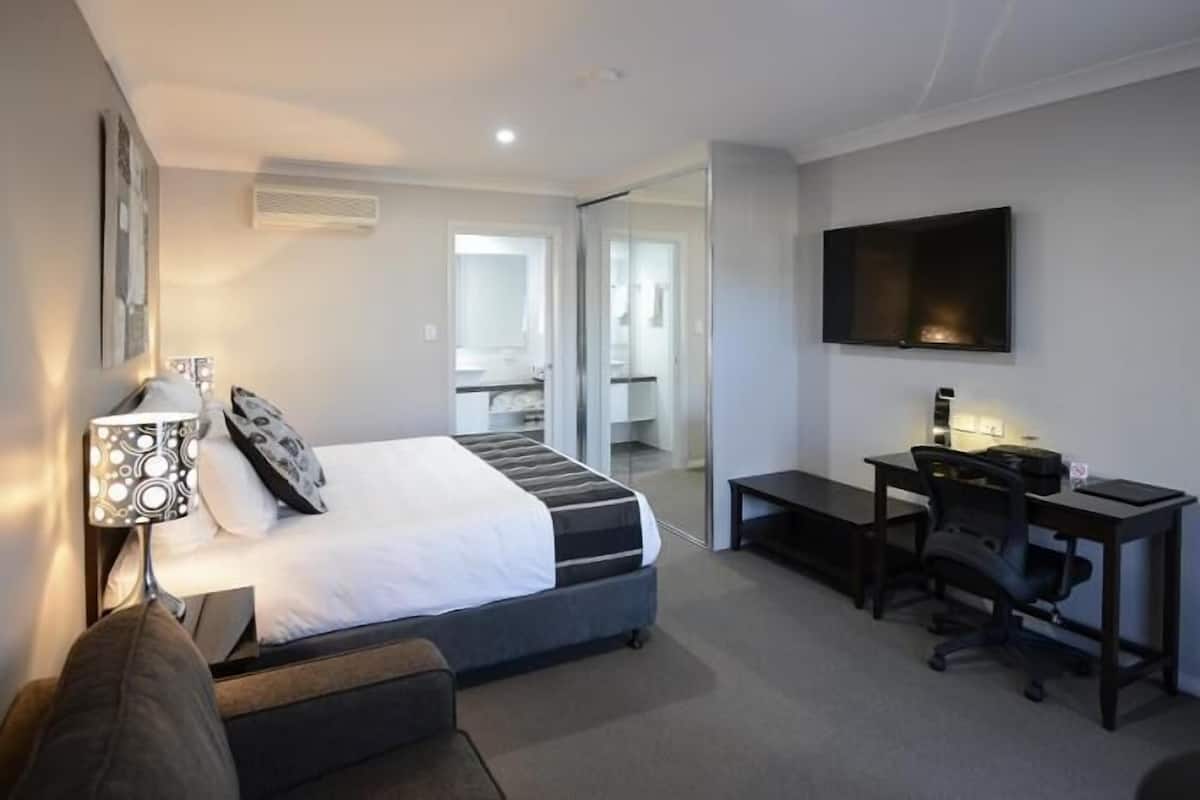 A hotel room with a bed, desk, chair, TV, and a bathroom visible through a glass door.