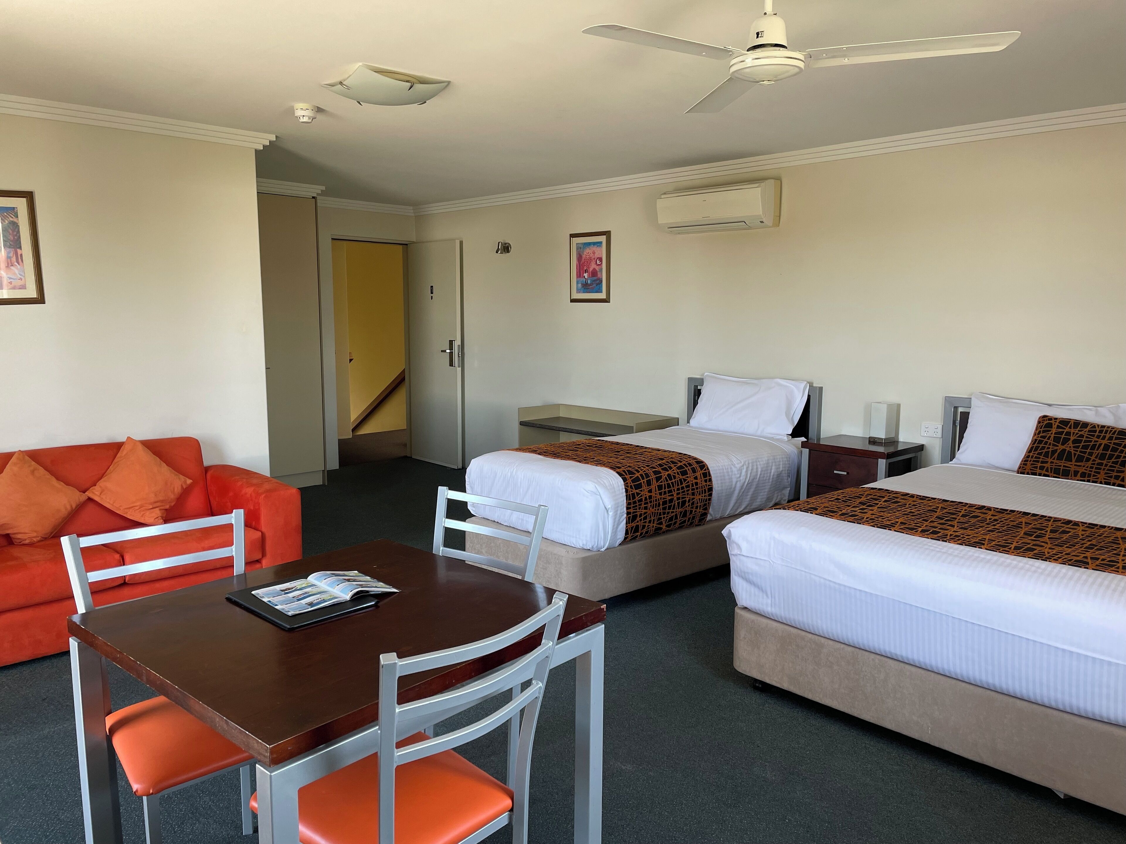 A hotel room with two beds, a dining table with a chair, a sofa, and a ceiling fan.