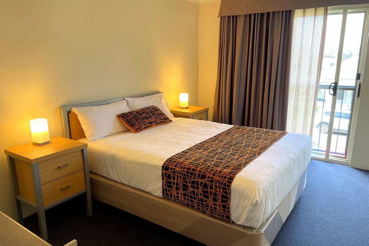 Habitación de hotel con una cama, dos mesitas de noche, una ventana con cortinas y vistas al exterior.