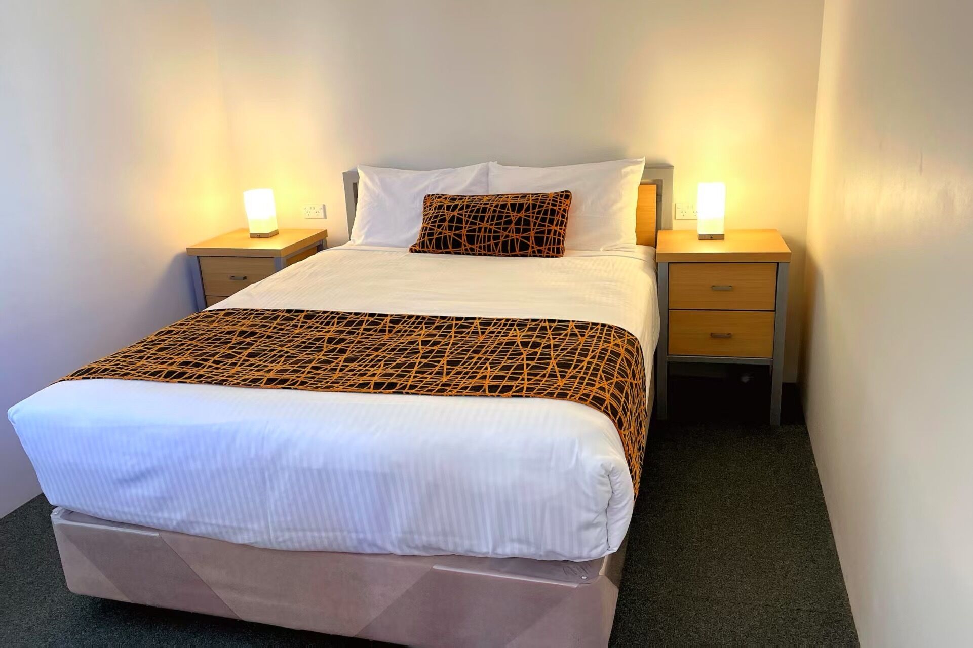 Un lit bien fait, recouvert d’une couvre-lit à motifs, deux tables de chevet avec des lampes et un tiroir.