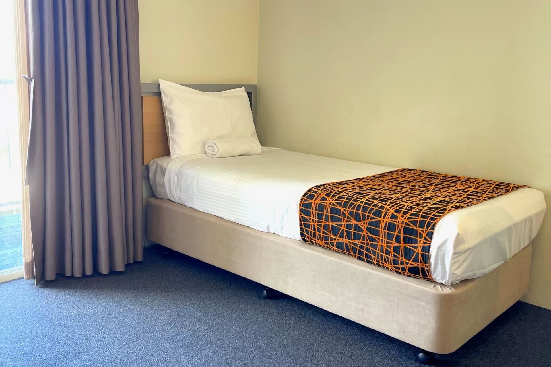 Una cama individual con colchón blanco y manta estampada, cabecera de madera y cortina gris.