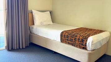 Una cama individual con colchón blanco y manta estampada, cabecera de madera y cortina gris.