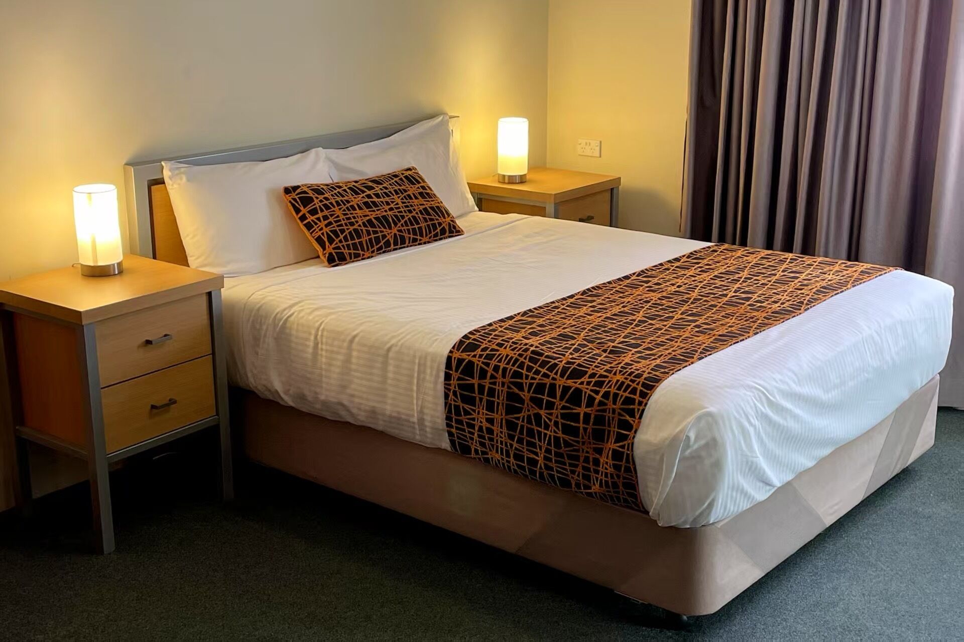 A hotel room with a bed, two bedside tables with lamps, a nightstand, a window with curtains, and a carpeted floor.