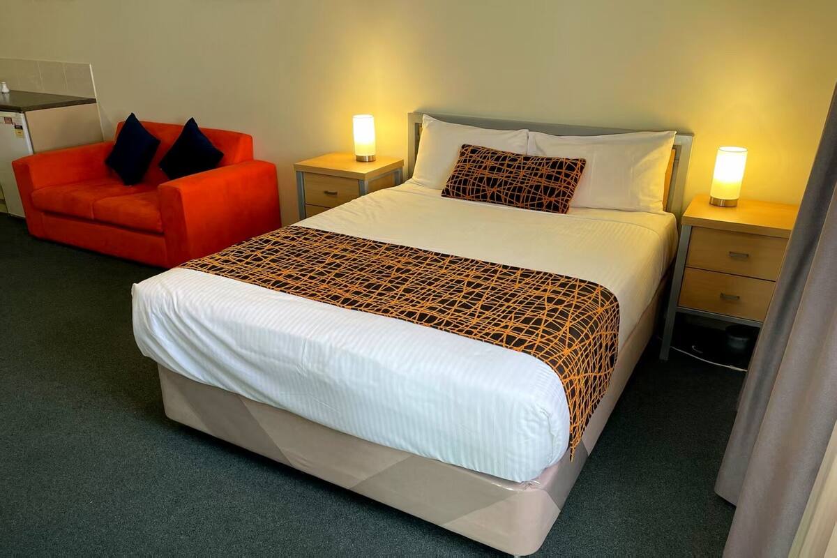 Una habitación de hotel con una cama grande, dos mesitas de noche con lámparas, un sillón rojo y un escritorio pequeño con una silla.