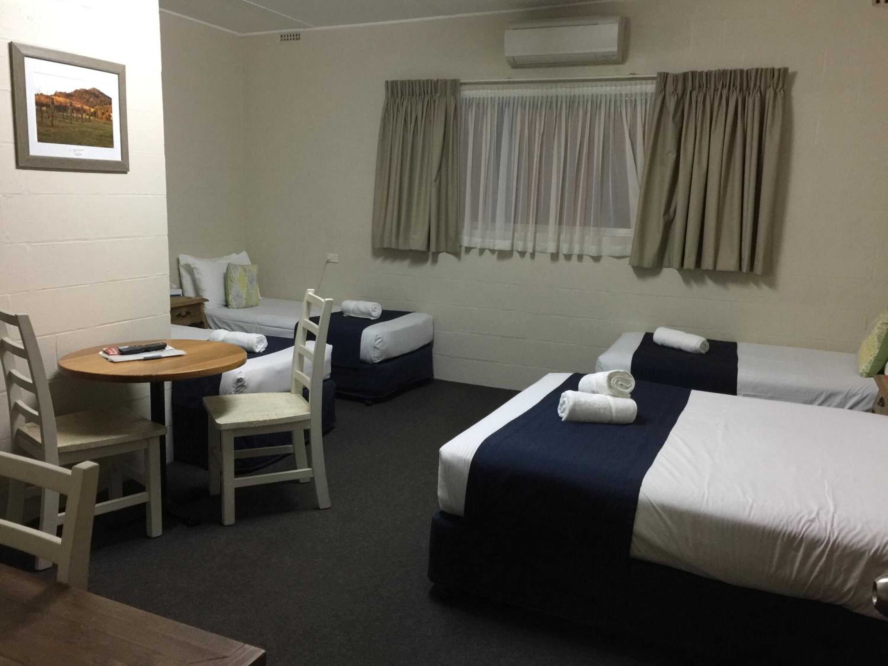 A hotel room with two single beds, a small table with a chair, and a single bed with a pillow and a blanket.