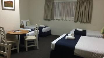 A hotel room with two single beds, a small table with a chair, and a single bed with a pillow and a blanket.