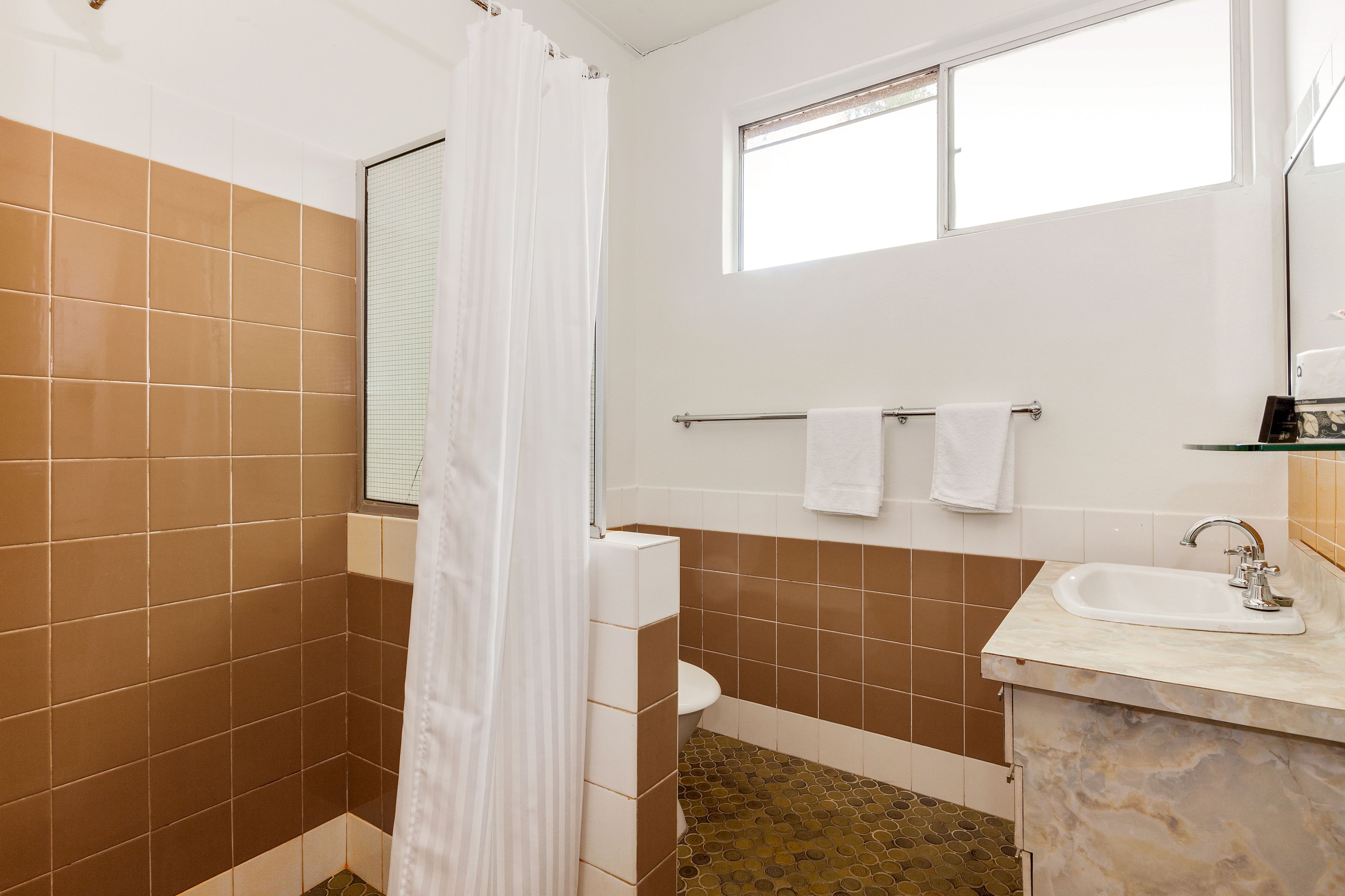 A bathroom with brown and white tiles, a white shower curtain, a window, a toilet, and a sink with a mirror.