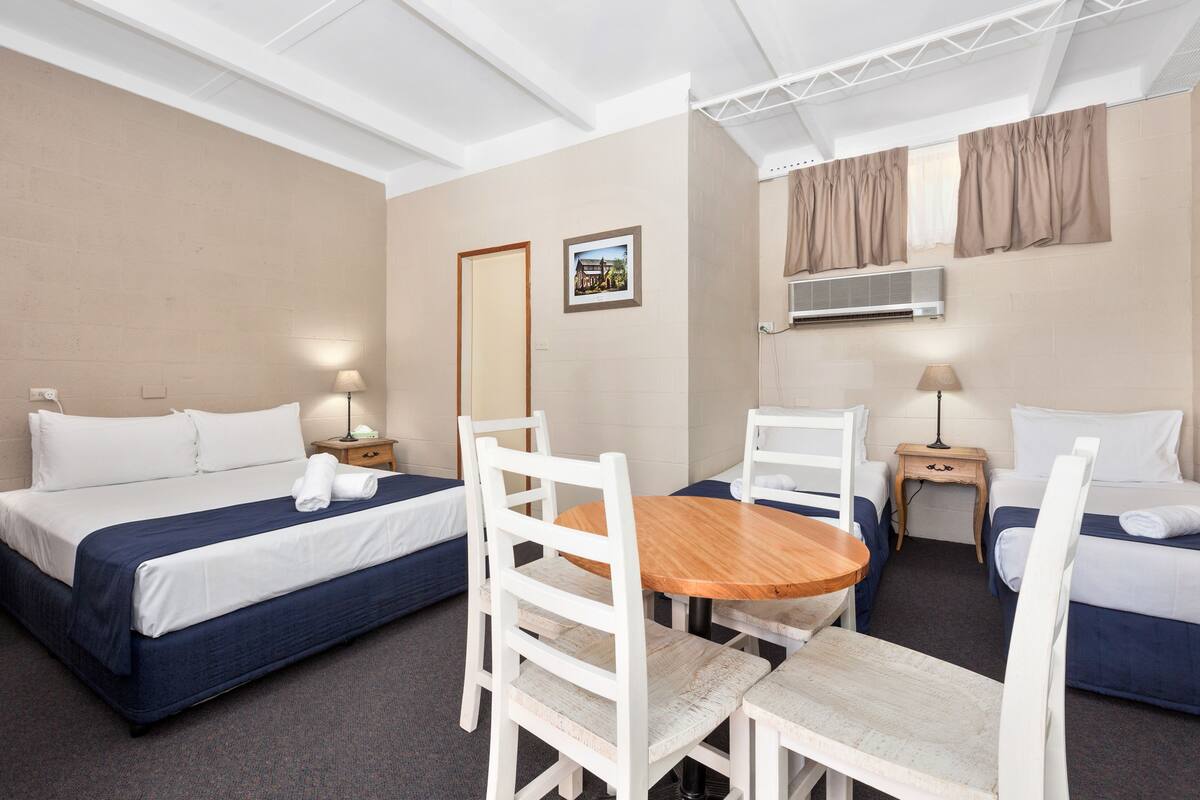 Quarto de hotel com duas camas, uma mesa de madeira, cadeiras brancas, um espelho e um quadro na parede.