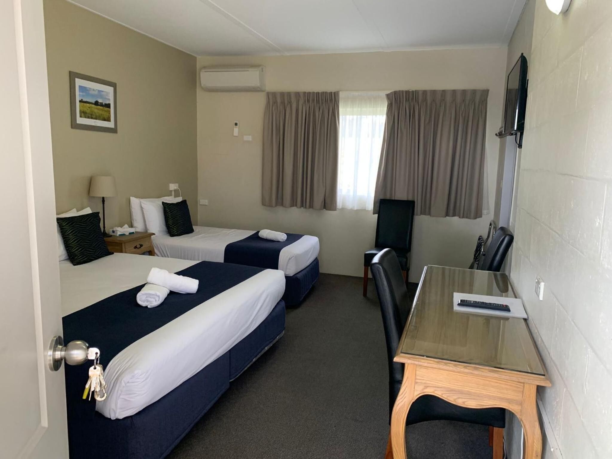 A hotel room with two beds, a desk, a chair, an air conditioning unit, a television, and a window with curtains.