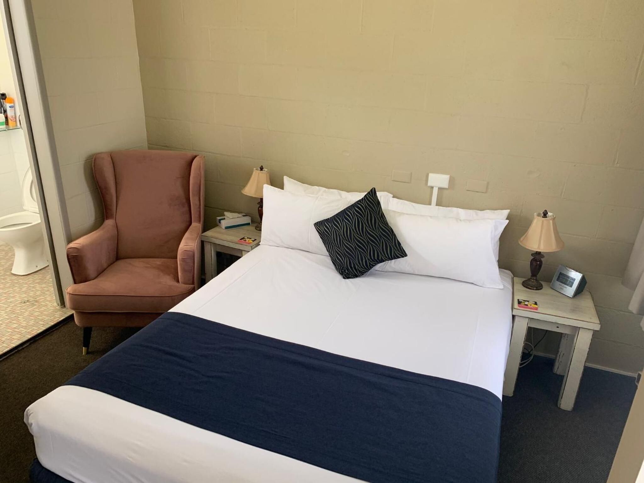 A hotel room with a bed, a chair, a nightstand, and a bedside table.